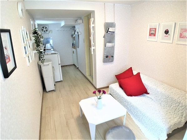 A small, cozy room with a white sofa, a small table, and a white cabinet. There are framed pictures on the wall and a plant on the table.