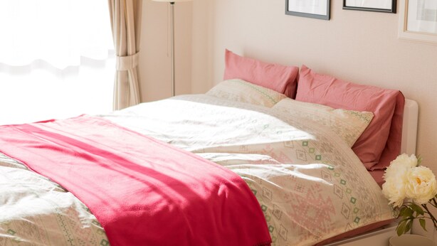 Una cama bien hecha con una manta rosa y almohadas, una lámpara de noche y un cuadro en la pared.