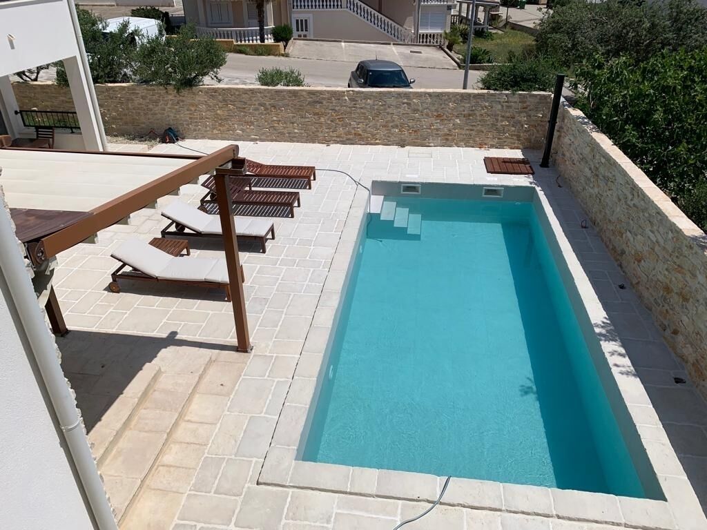 A rectangular swimming pool with a tiled deck, surrounded by a stone wall and a wooden pergola with lounge chairs.