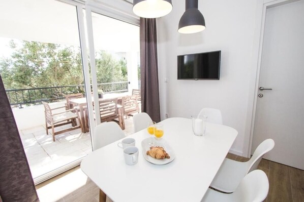 Un coin repas moderne avec une table blanche, des chaises et une table dressée pour le petit-déjeuner, comprenant un croissant, du café et du jus d'orange.