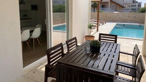 A patio with a table and chairs, a potted plant, and a view of a swimming pool and buildings in the background.