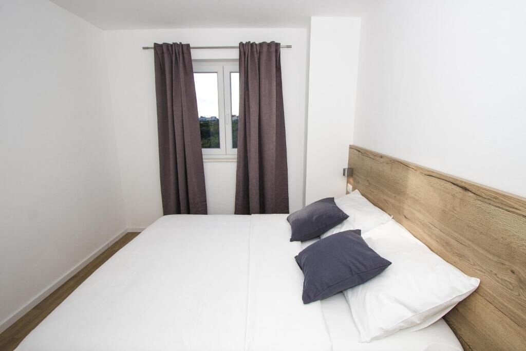 Een slaapkamer met een enkel bed, twee kussens, een houten hoofdbord en een raam met gordijnen.