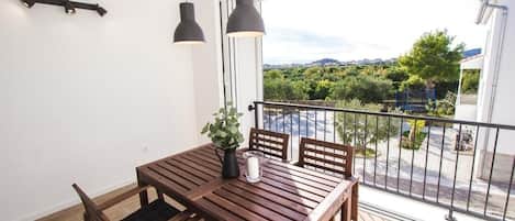 A balcony with a wooden table and chairs, a potted plant, and a view of greenery and buildings.