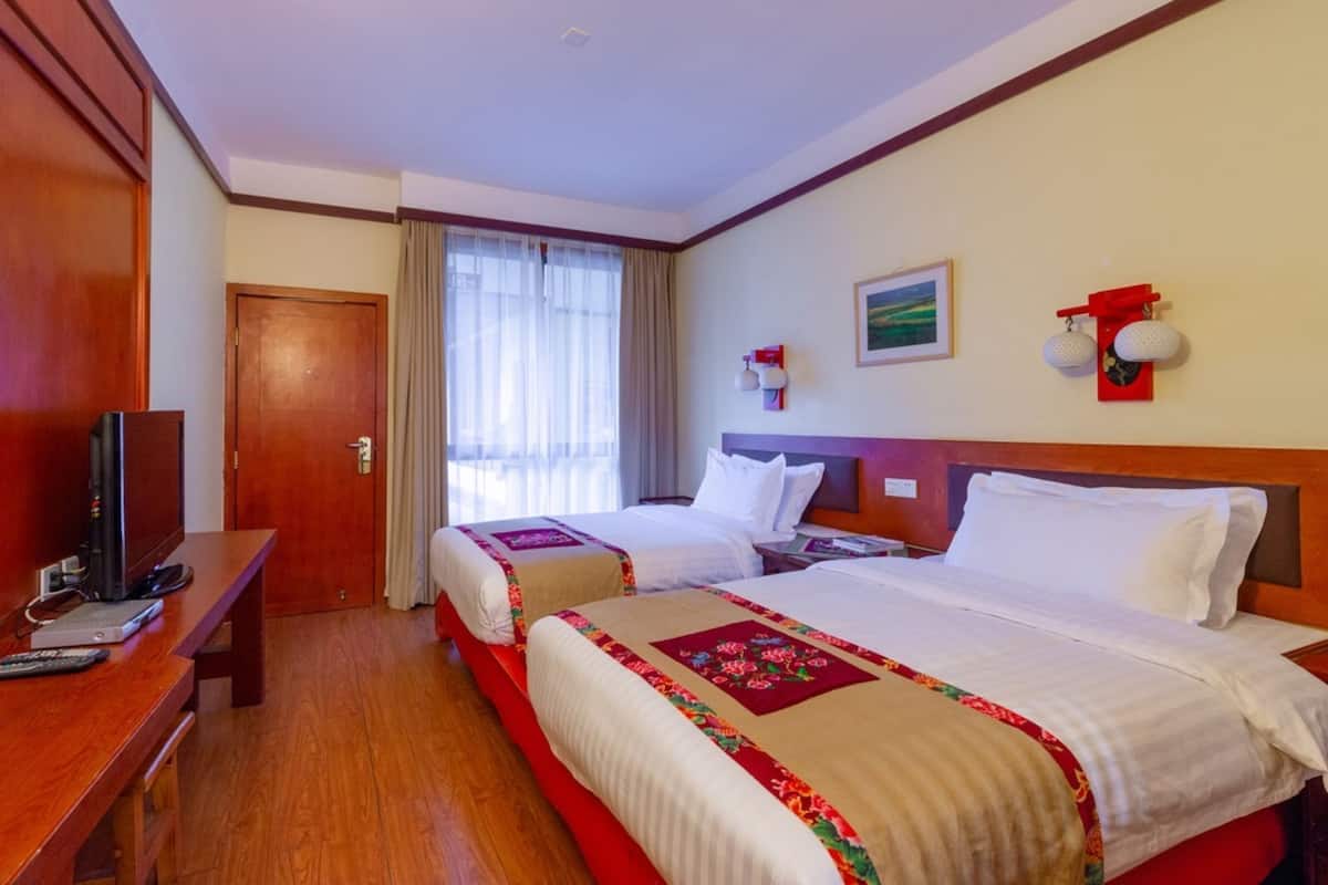A hotel room with two beds, a desk with a TV, a wooden wardrobe, and a window with curtains.