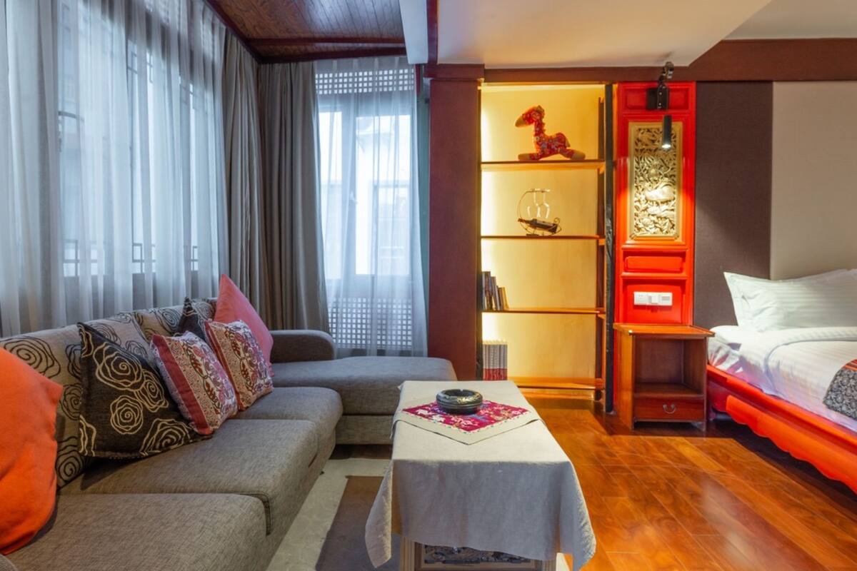 A modern hotel room with a sofa, a small table, a bed, and a decorative shelf with books and a statue.