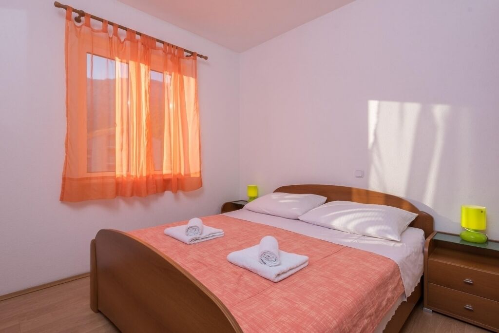 A neatly made bed with a pink bedspread and two white pillows, a wooden headboard, a nightstand with a green lamp, and an orange sheer curtain over a window.