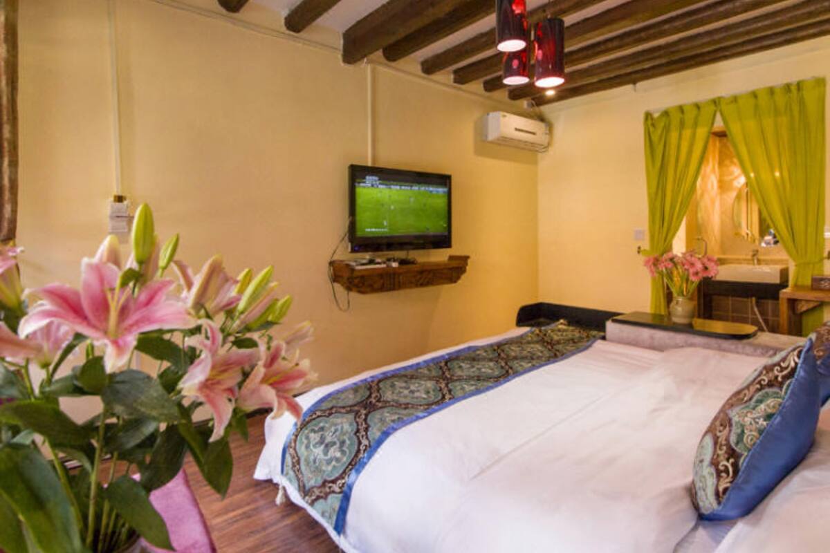 A hotel room with a bed, a television, a small shelf, a window with curtains, and a vase with flowers.