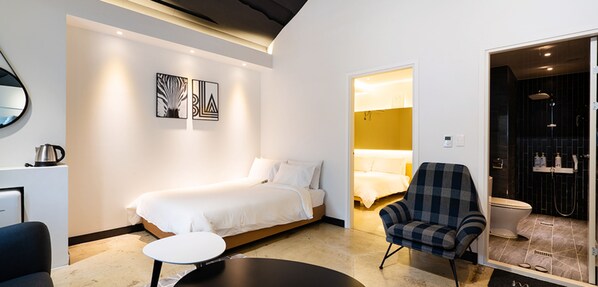 Habitación de hotel moderna con cama, una silla, una mesita y un baño visible a través de una puerta abierta.