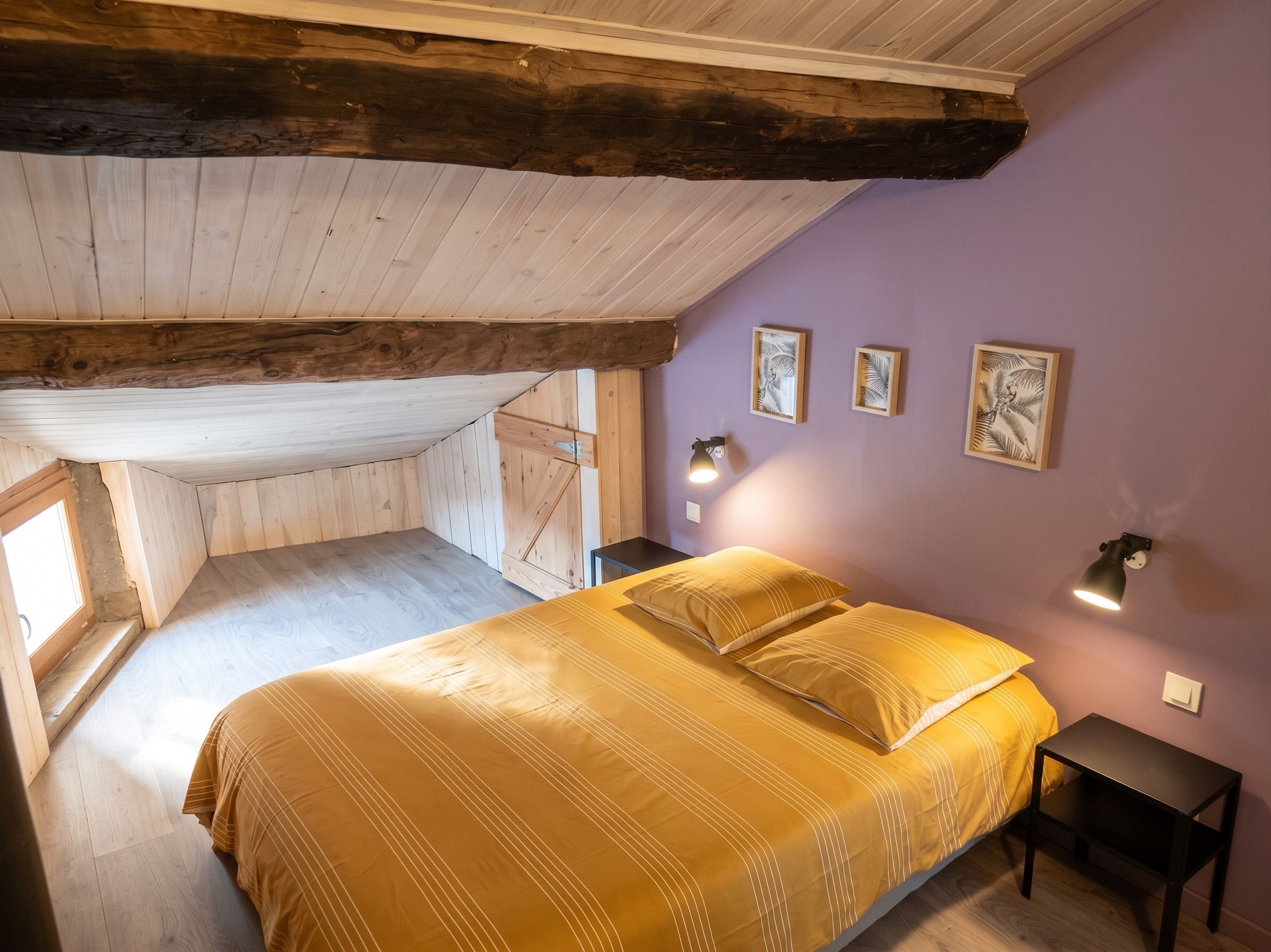 Une chambre avec un plafond en pente, des poutres en bois, un lit avec une literie jaune, deux tables de chevet et des lampes fixées au mur.
