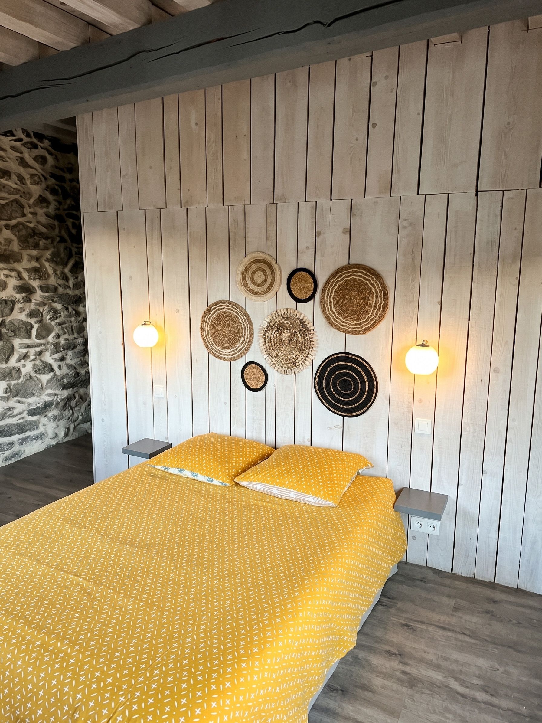 A bed with a yellow patterned bedspread, a stone wall to the left, and a wooden wall with wall art and wall sconces.