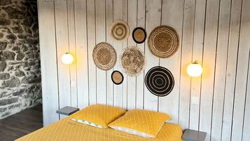 A bed with a yellow patterned bedspread, a stone wall to the left, and a wooden wall with wall art and wall sconces.