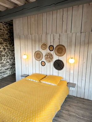 A bed with a yellow patterned bedspread, a stone wall to the left, and a wooden wall with wall art and wall sconces.