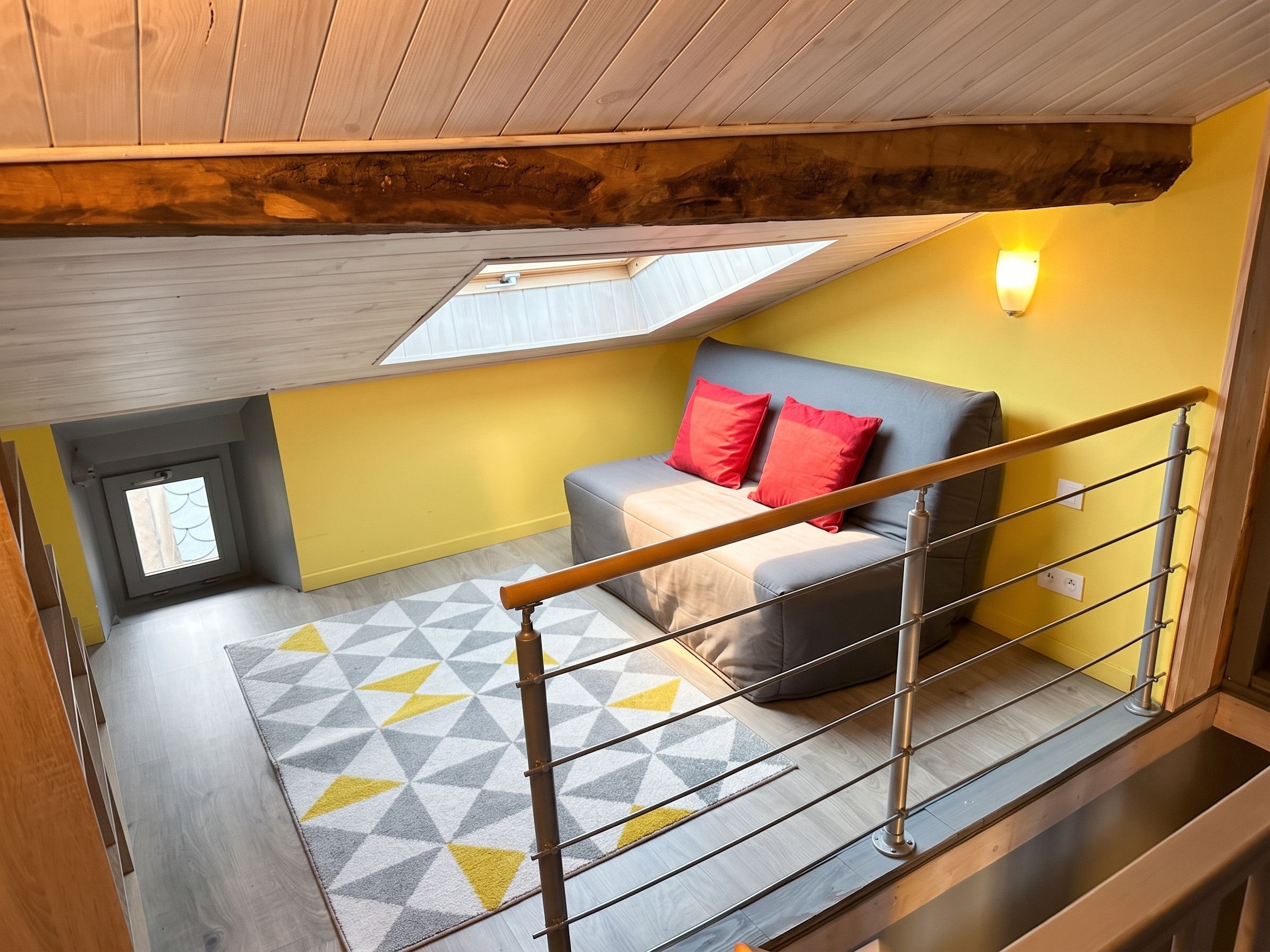 Un dormitorio con techo inclinado, un tragaluz, una cama con almohadas rojas, un tapiz con estampado geométrico y una barandilla de metal.