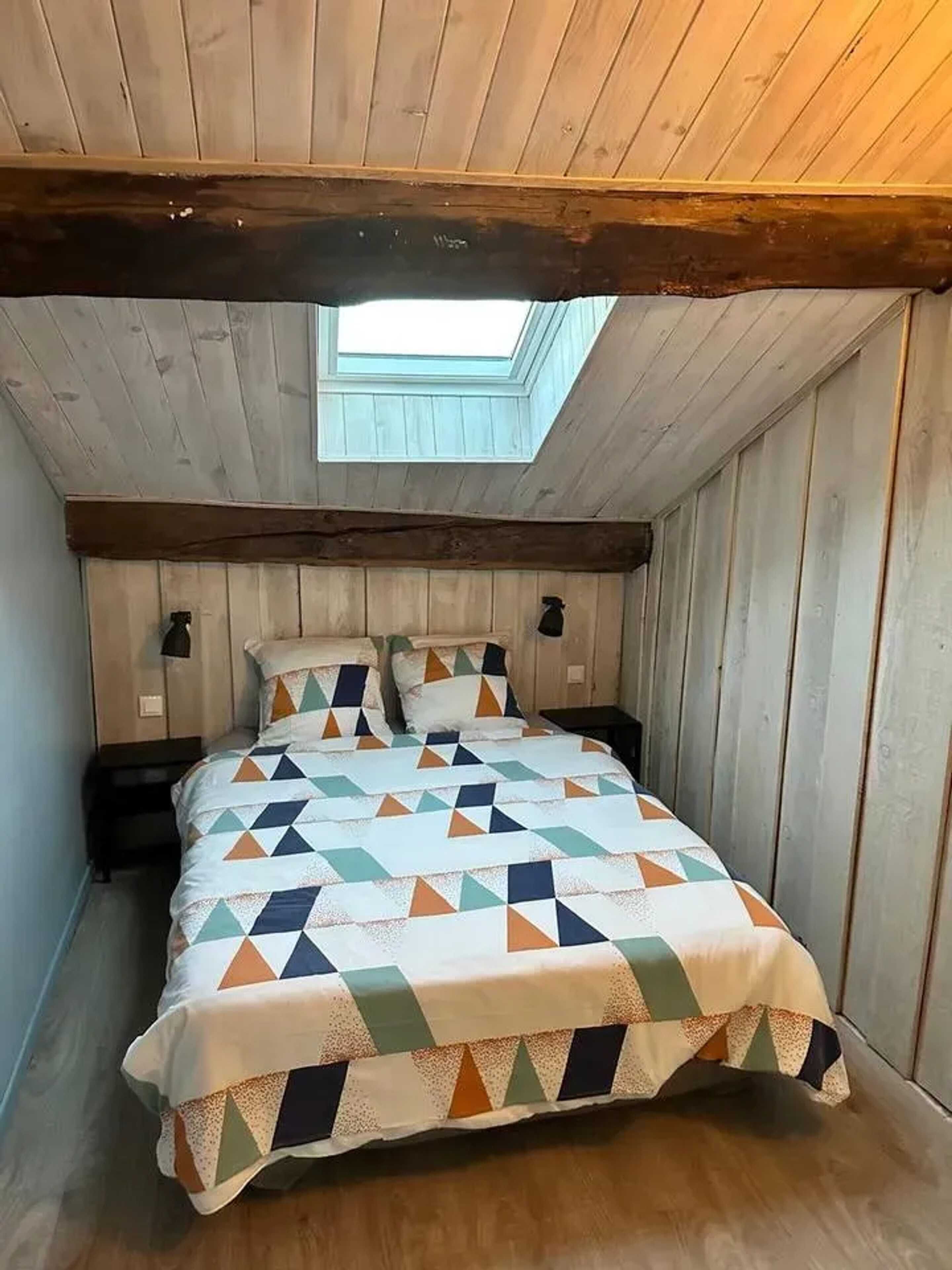 Une petite chambre mansardée, chaleureuse et accueillante, avec un plafond en pente, une lucarne et un lit recouvert d’une courtepointe à motifs géométriques.