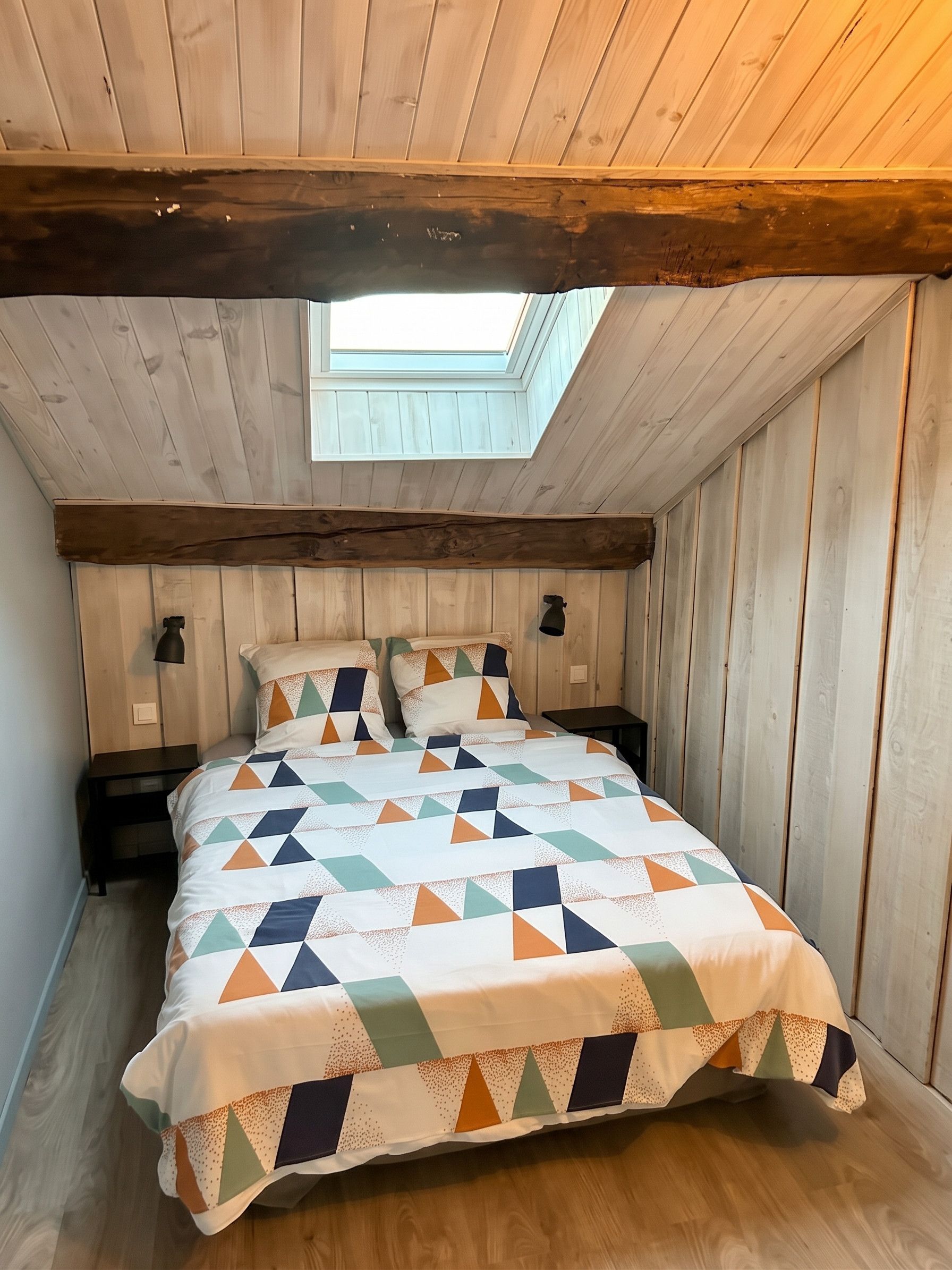 A bedroom with a sloped ceiling, a skylight, a bed with a patterned bedspread, two bedside lamps, and a wooden floor.