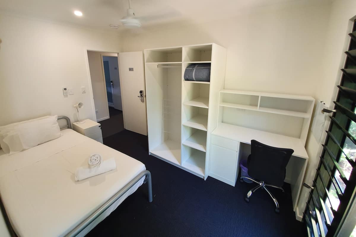 Een hotelkamer met een bed, een bureau, een stoel, een kledingkast en een plafondventilator.