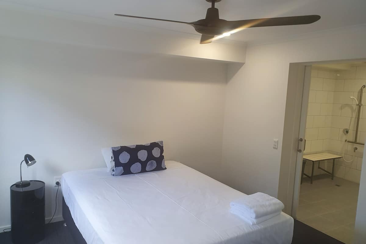 Een hotelkamer met een bed, een plafondventilator, een bedlampje en een zichtbare badkamer door een geopende deur.
