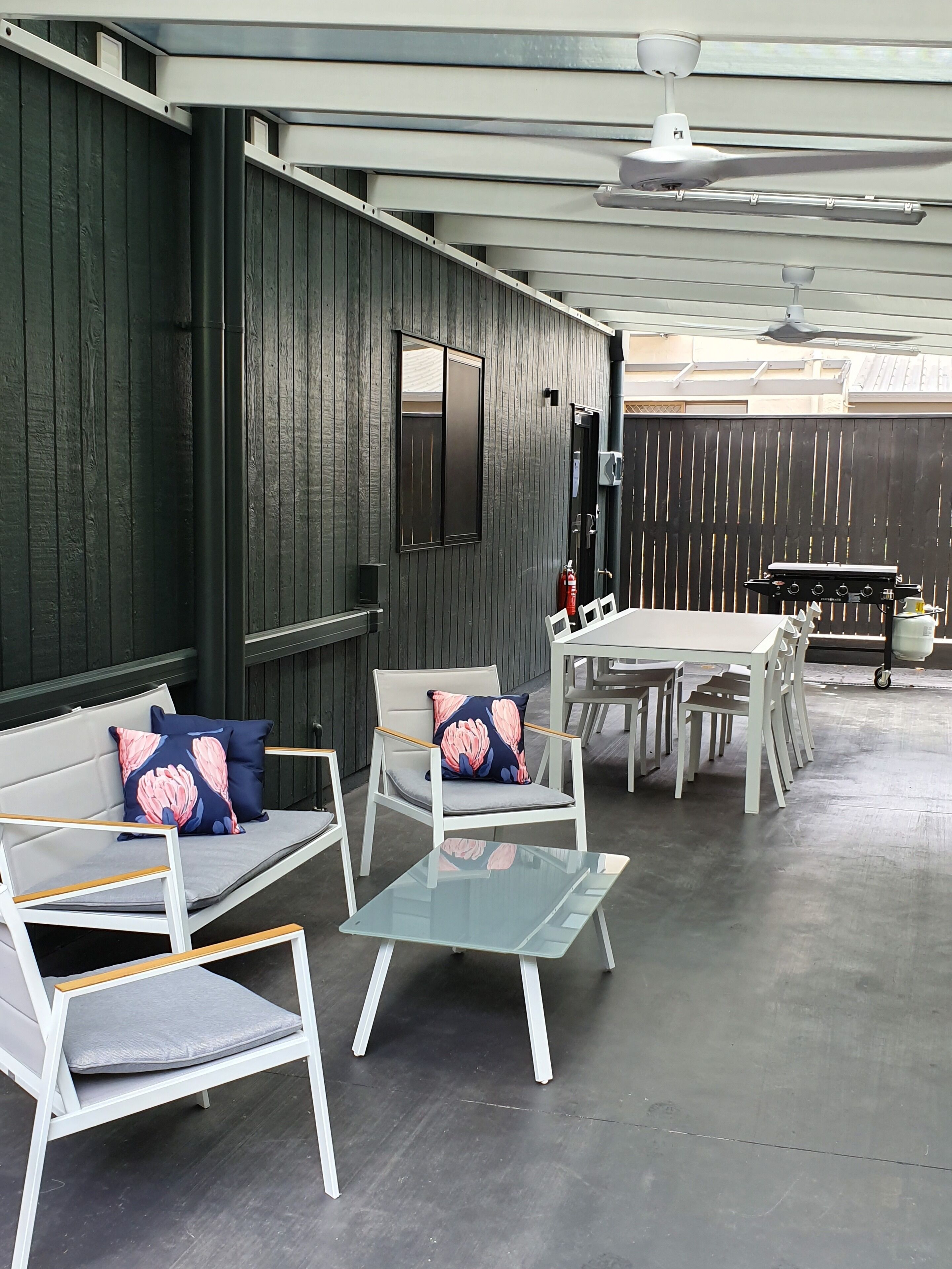 An outdoor seating area with white furniture and a glass table, surrounded by dark wooden walls and a ceiling fan.