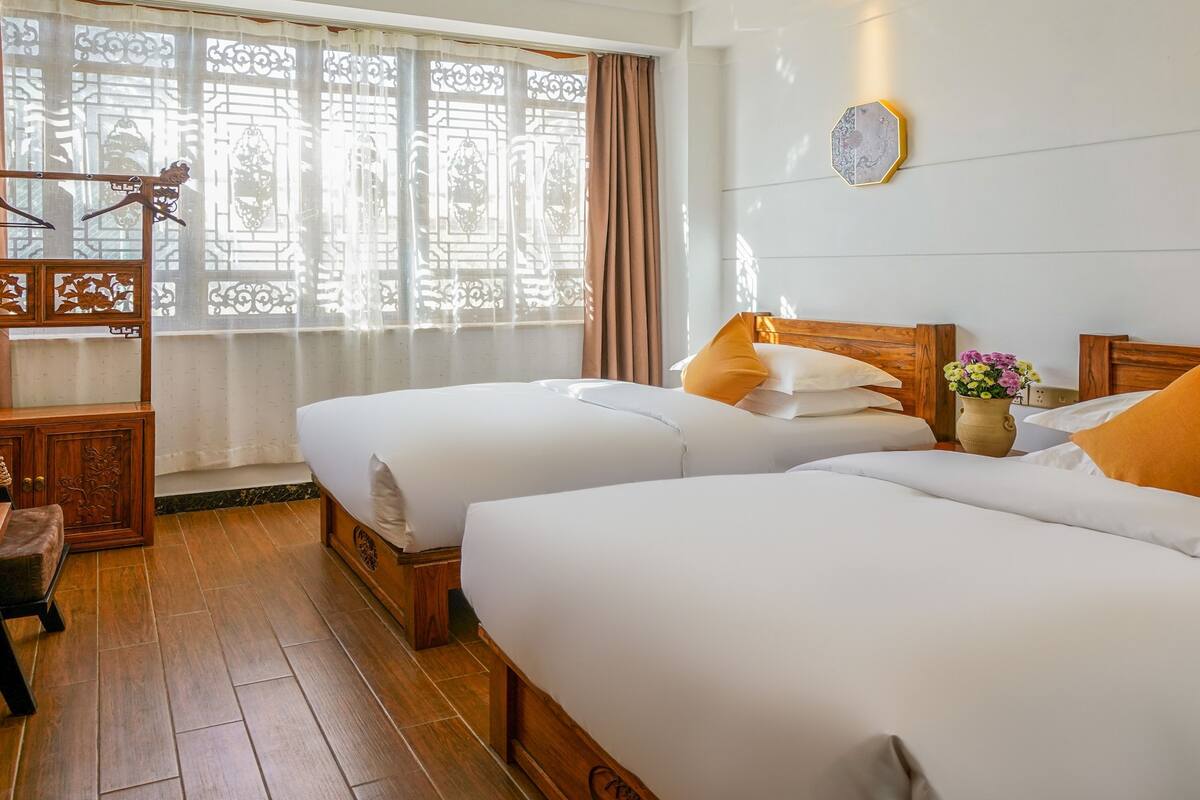 Habitación de hotel con dos camas, cabecera de madera, ventana decorativa con cortina y lámpara fijada a la pared.