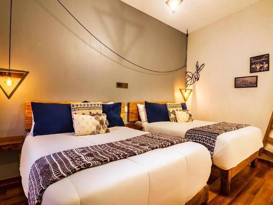 A hotel room with two beds, each with patterned bedding and pillows. There are wall-mounted lights and a wooden ladder.