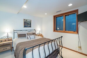 Un dormitorio con cama, mesitas de noche, lámpara, ventana con cortinas y televisión montada en la pared.