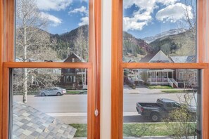 Vista a través de una ventana con marco de madera, mostrando una calle residencial con casas, coches estacionados y un paisaje montañoso al fondo.