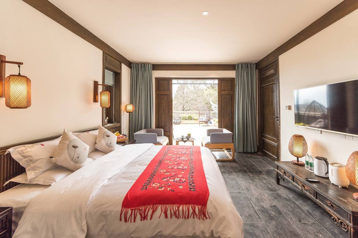 A hotel room with a large bed, a TV, a desk with a lamp, and a view of the outdoors.