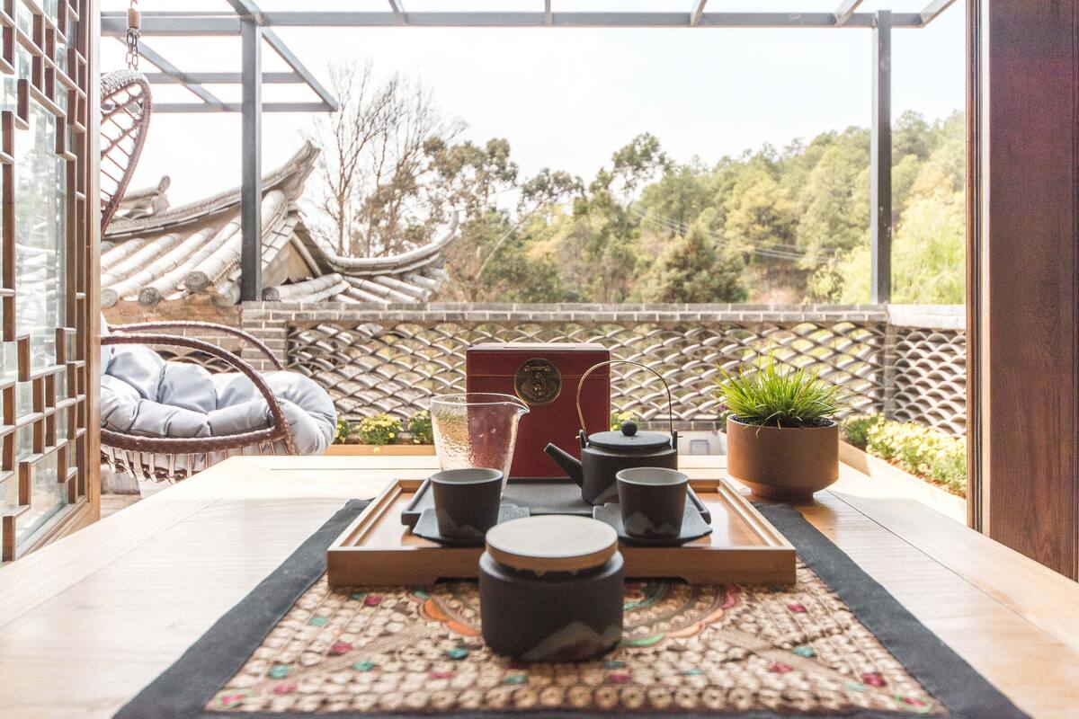 A traditional tea set on a tray, a wicker chair, and a potted plant on a balcony with a view of traditional architecture and trees.