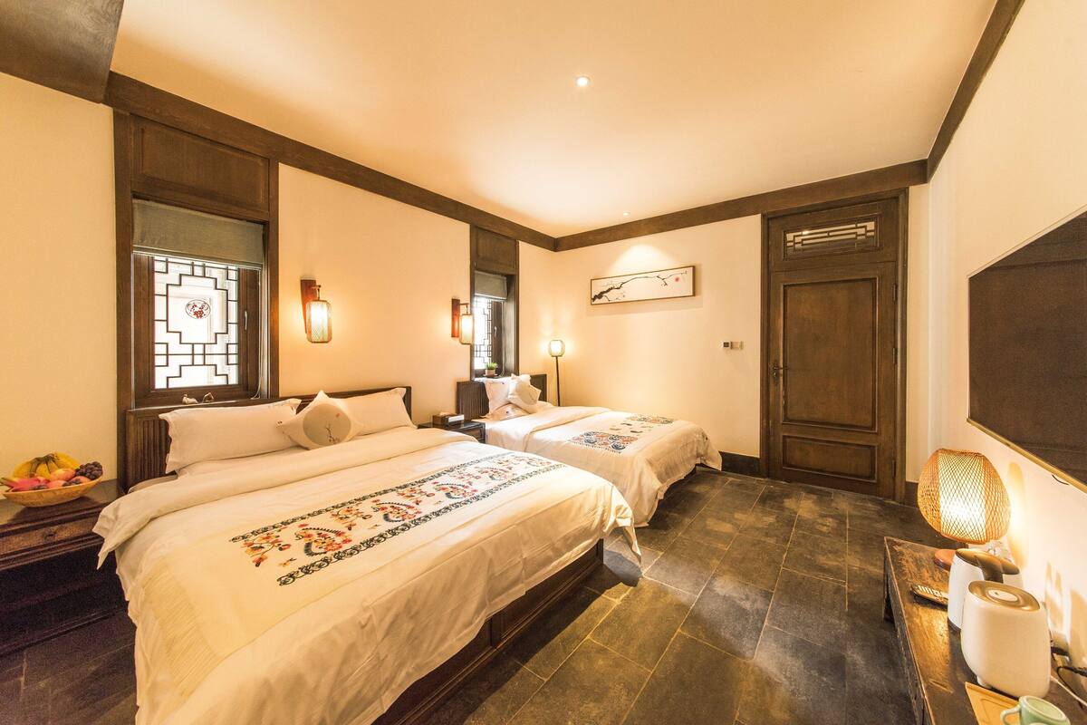 A hotel room with two beds, a wooden floor, a large window with a decorative glass design, and a wall-mounted lamp.