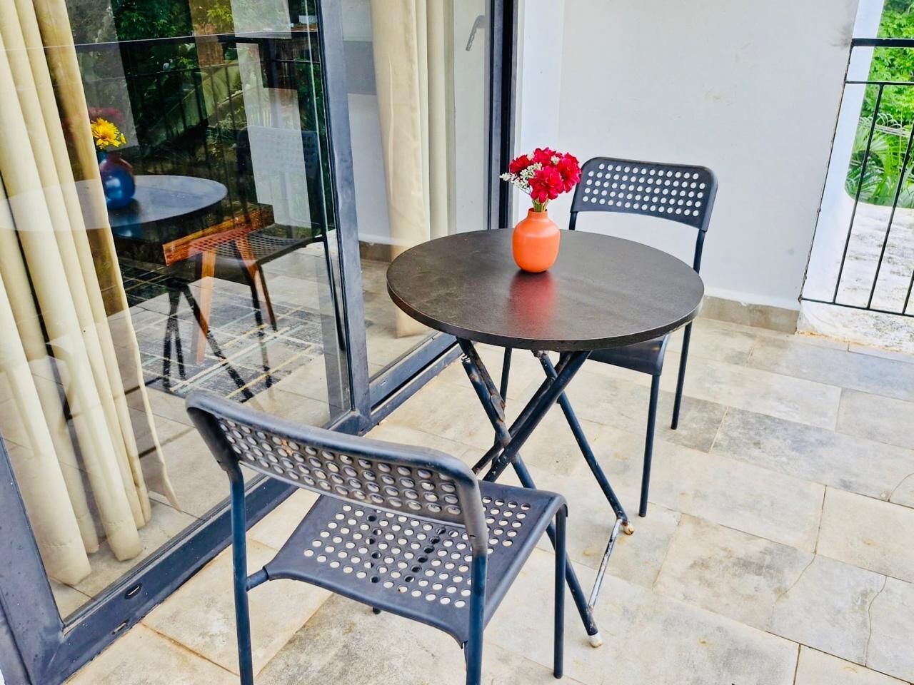 An outdoor patio with a round table and two chairs, a vase with flowers, and a glass door leading to a garden.