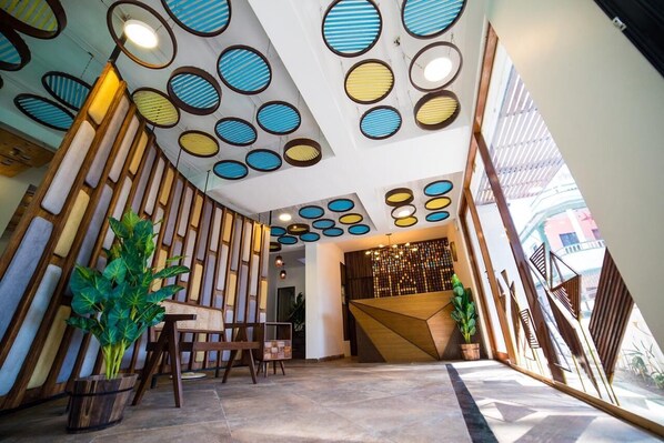 A modern reception area with a geometric wooden wall, a reception desk, and a seating area with a plant. - Half Hotel (Candolim)