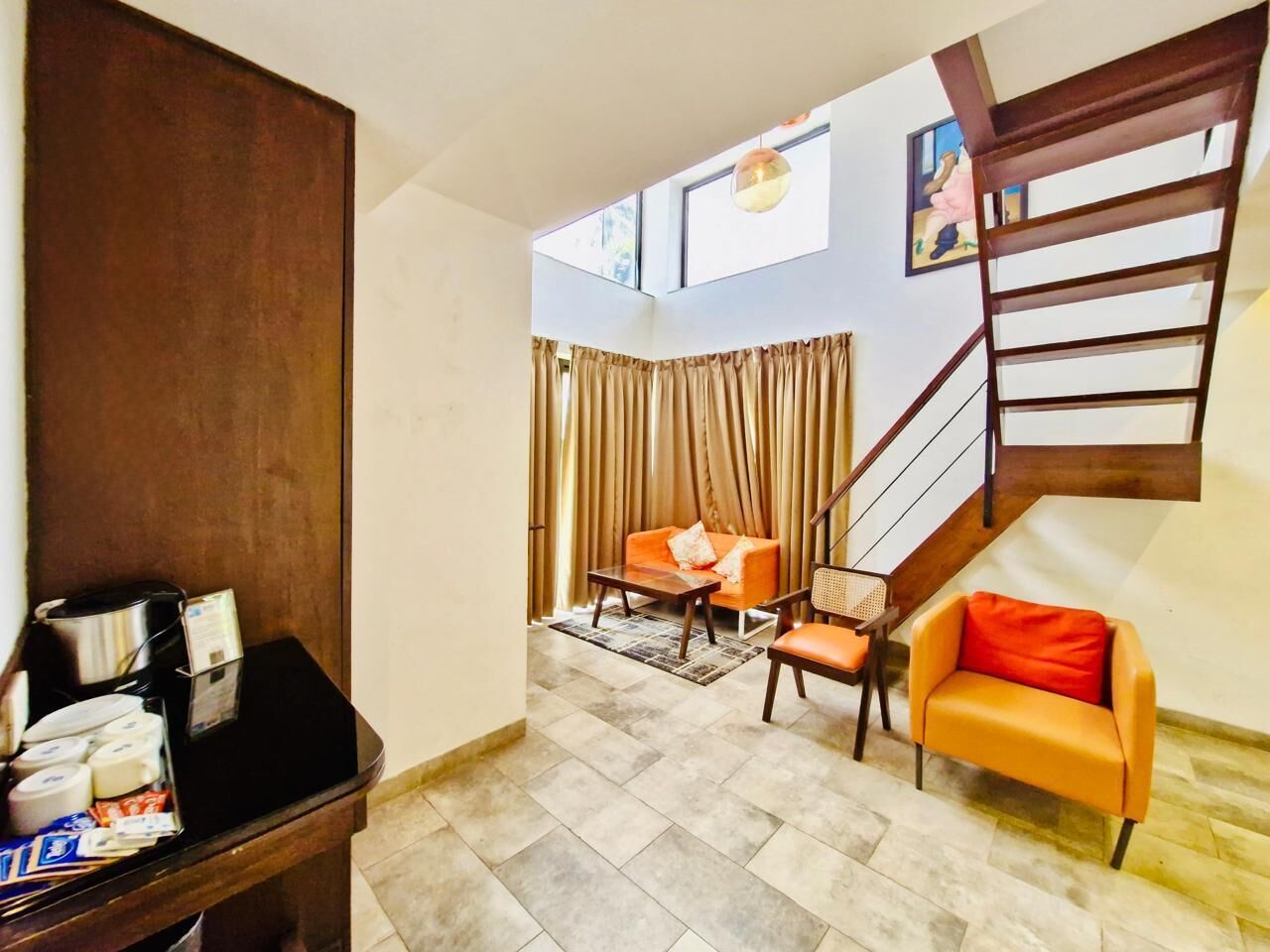 Una sala de estar con sofá, sillón, mesa de centro y una pequeña mesa con objetos encima. Se ve una escalera con barandilla y una ventana con cortinas.