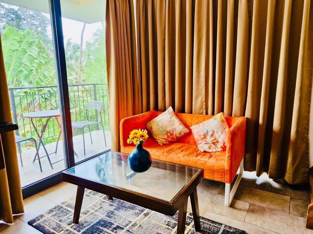 Una sala de estar con una mesa de centro de vidrio, un sofá naranja con cojines estampados y un florero con flores amarillas.