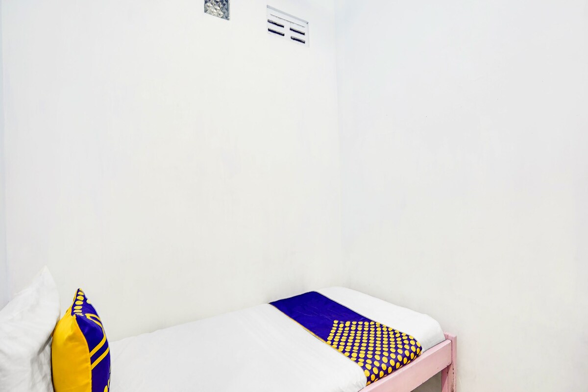 Una cama individual con colchón blanco, almohada de diseño azul y amarillo, y cabecera rosa en una habitación con paredes blancas y suelo de baldosas.