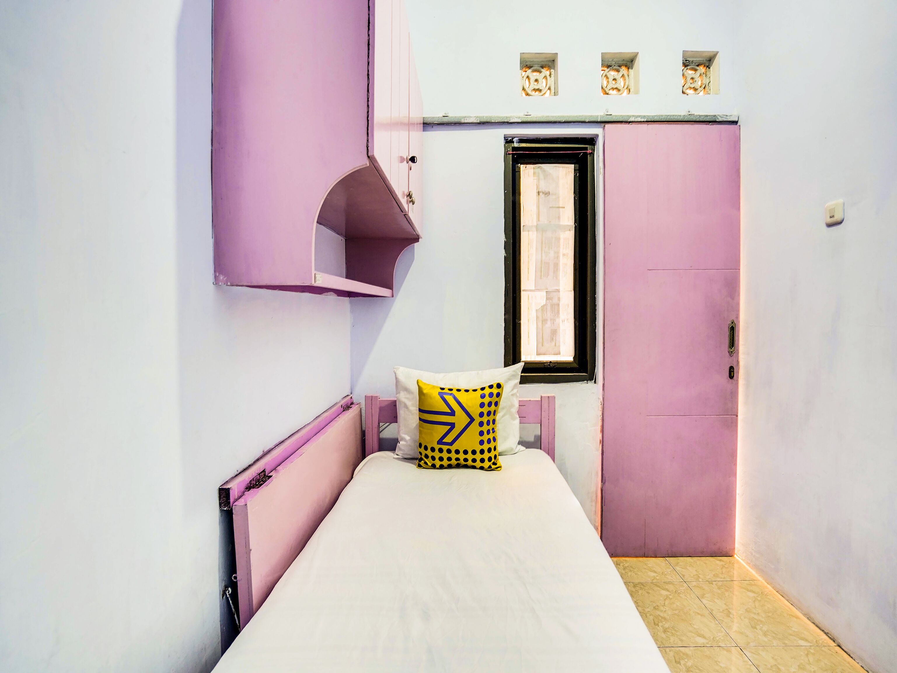 Una habitación pequeña con cama individual, decorada en tonos rosa y blanco, ventana con vistas y una puerta con un pequeño ventanal encima.