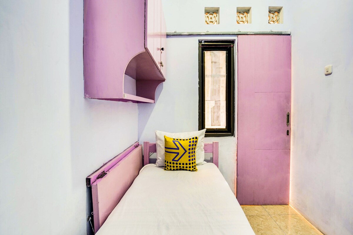 Habitació petita amb un llit individual, decorada amb tons rosa i blancs, una finestra amb vistes i una porta amb un petit finestró a la part superior.