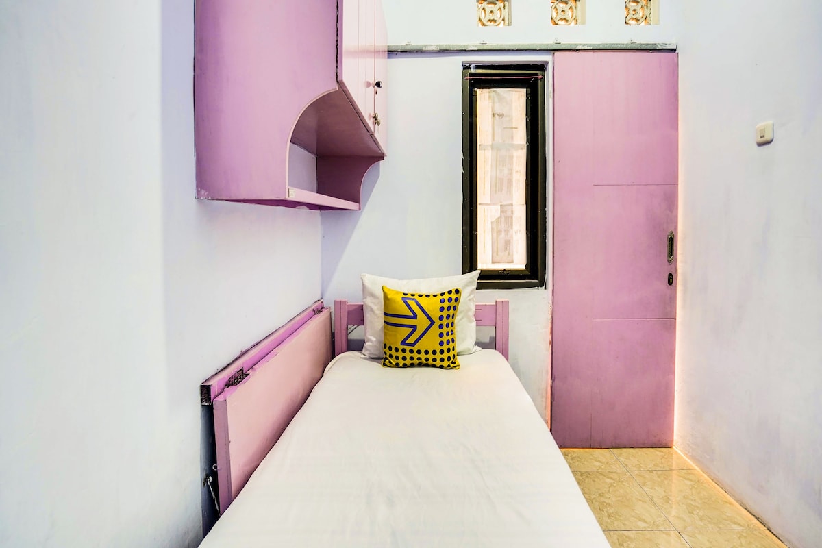 Una habitación pequeña con cama individual, decorada en tonos rosa y blanco, ventana con vistas y una puerta con un pequeño ventanal encima.