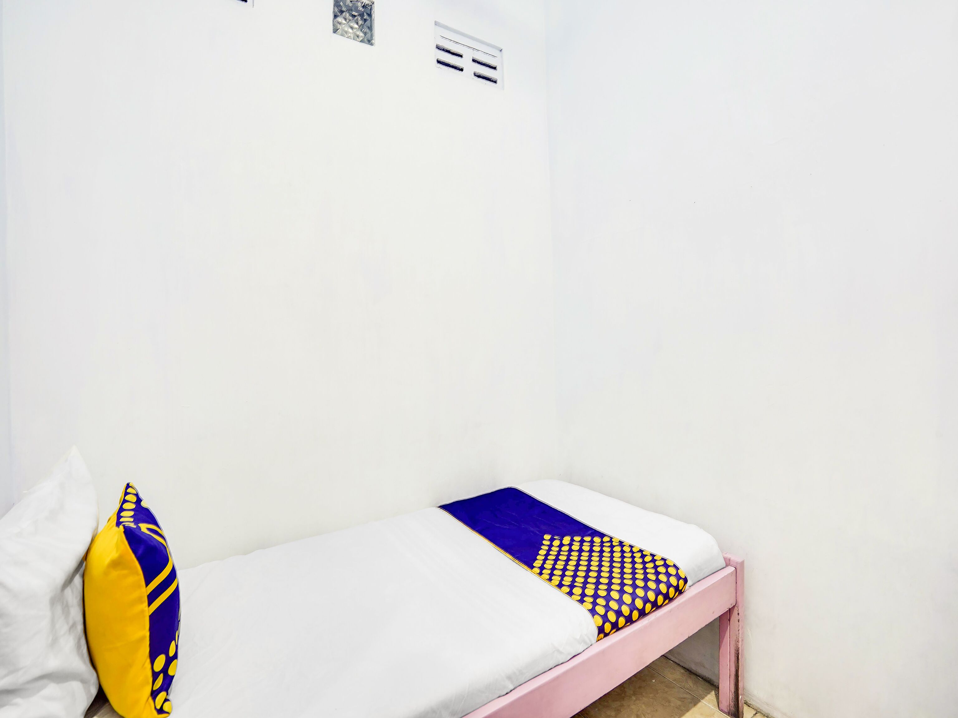 Una cama individual con colchón blanco, almohada de diseño azul y amarillo, y cabecera rosa en una habitación con paredes blancas y suelo de baldosas.