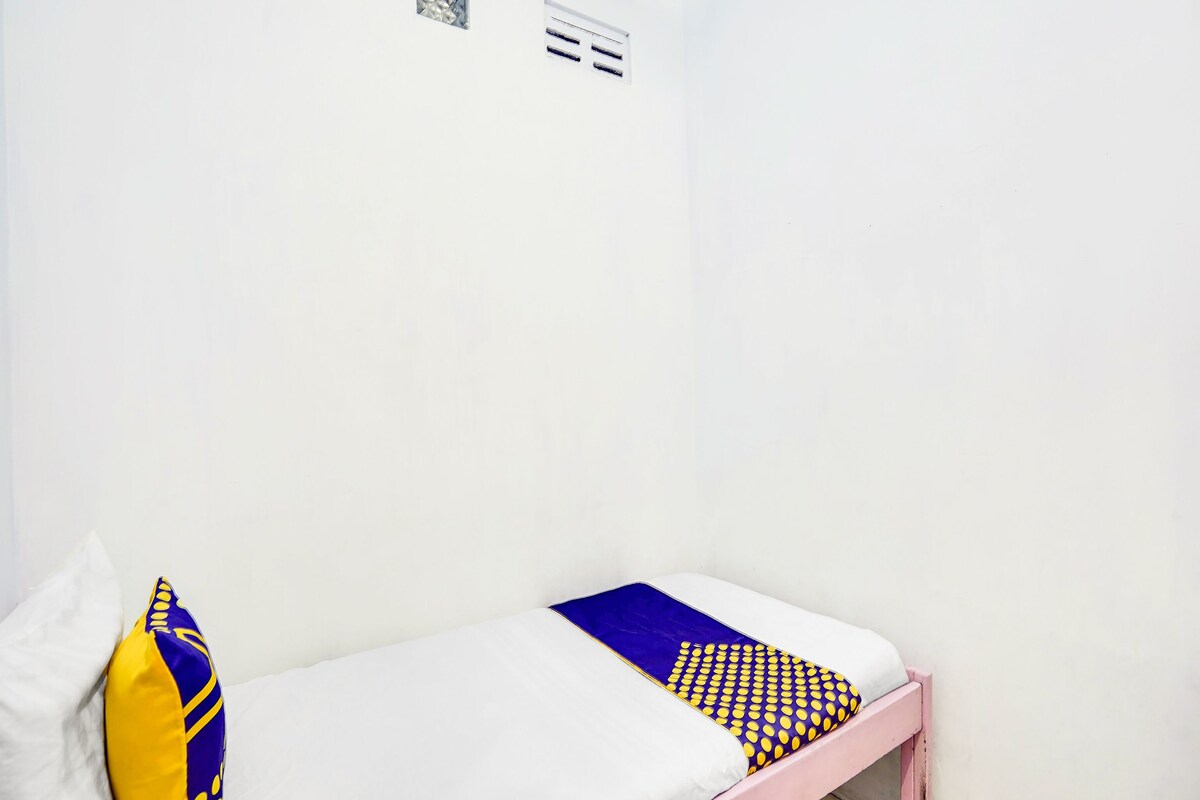 Un llit individual amb matalàssi blanc, un coixí amb dibuixos blaus i grocs i un capçal de llit rosa en una habitació amb parets blanques i terra de rajola.