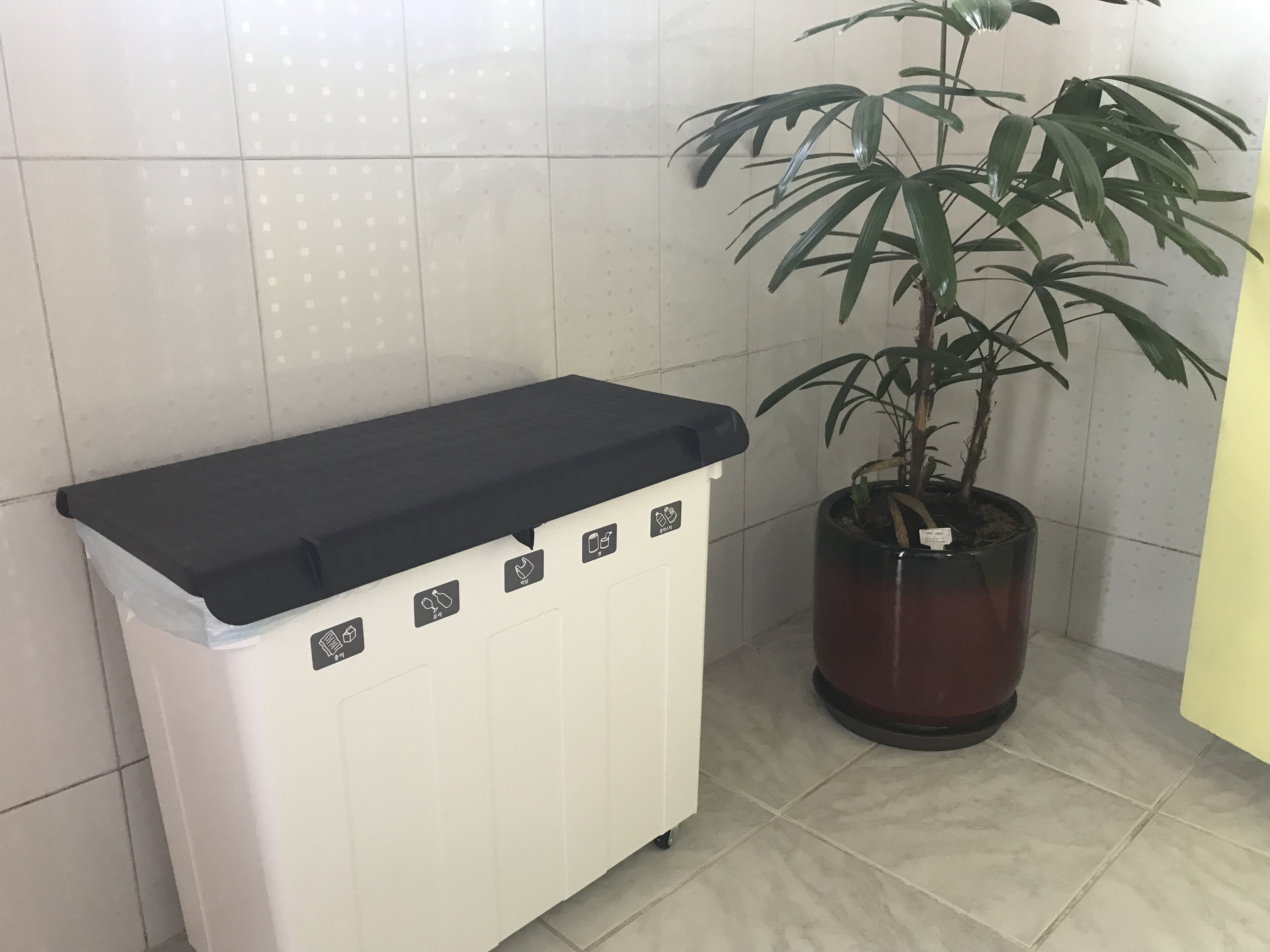 A white utility cart with a black top, a potted plant, and a tiled wall.