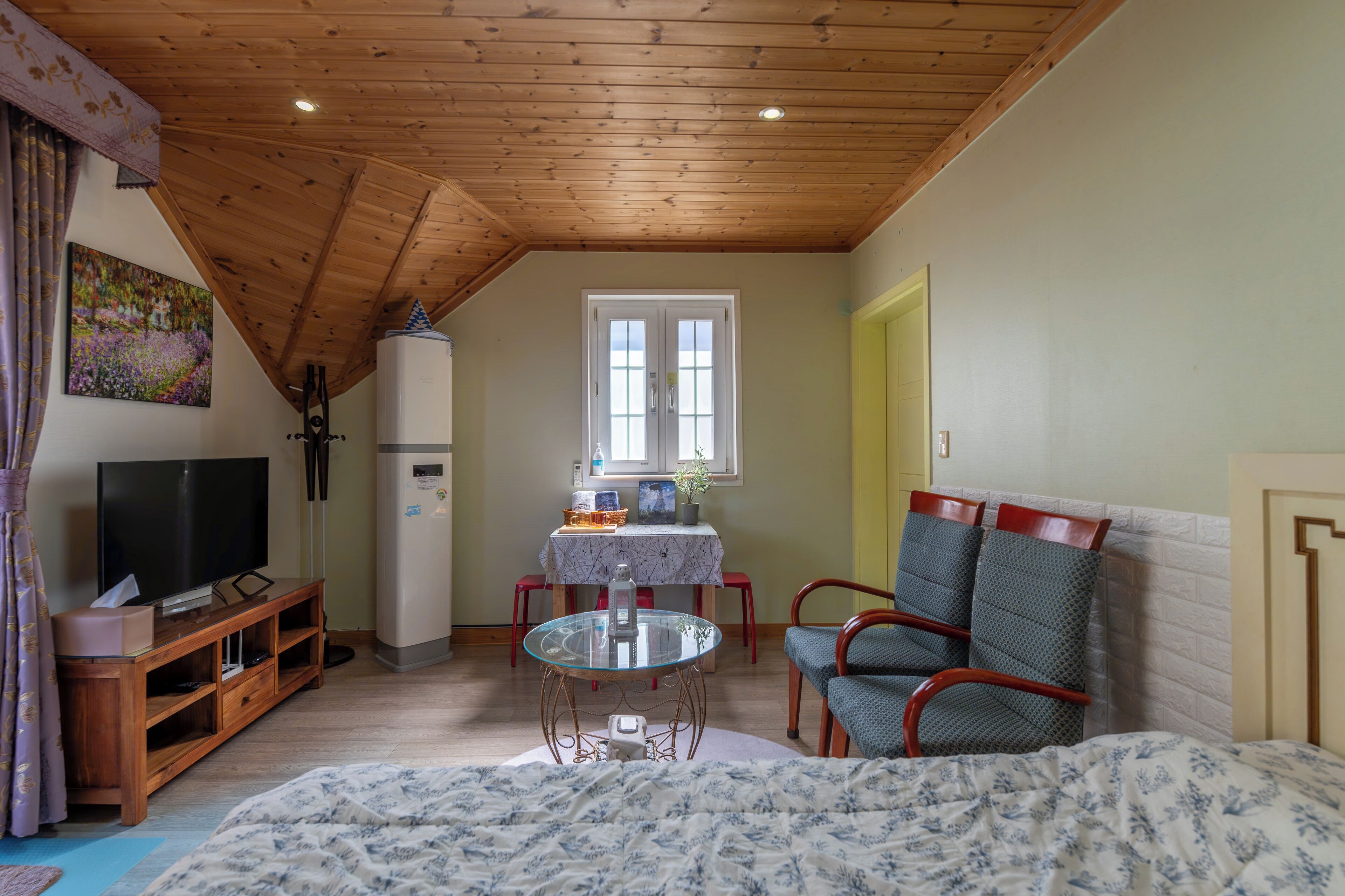 A cozy room with a wooden ceiling, a television, a refrigerator, a dining table with chairs, and a bed with a floral bedspread.