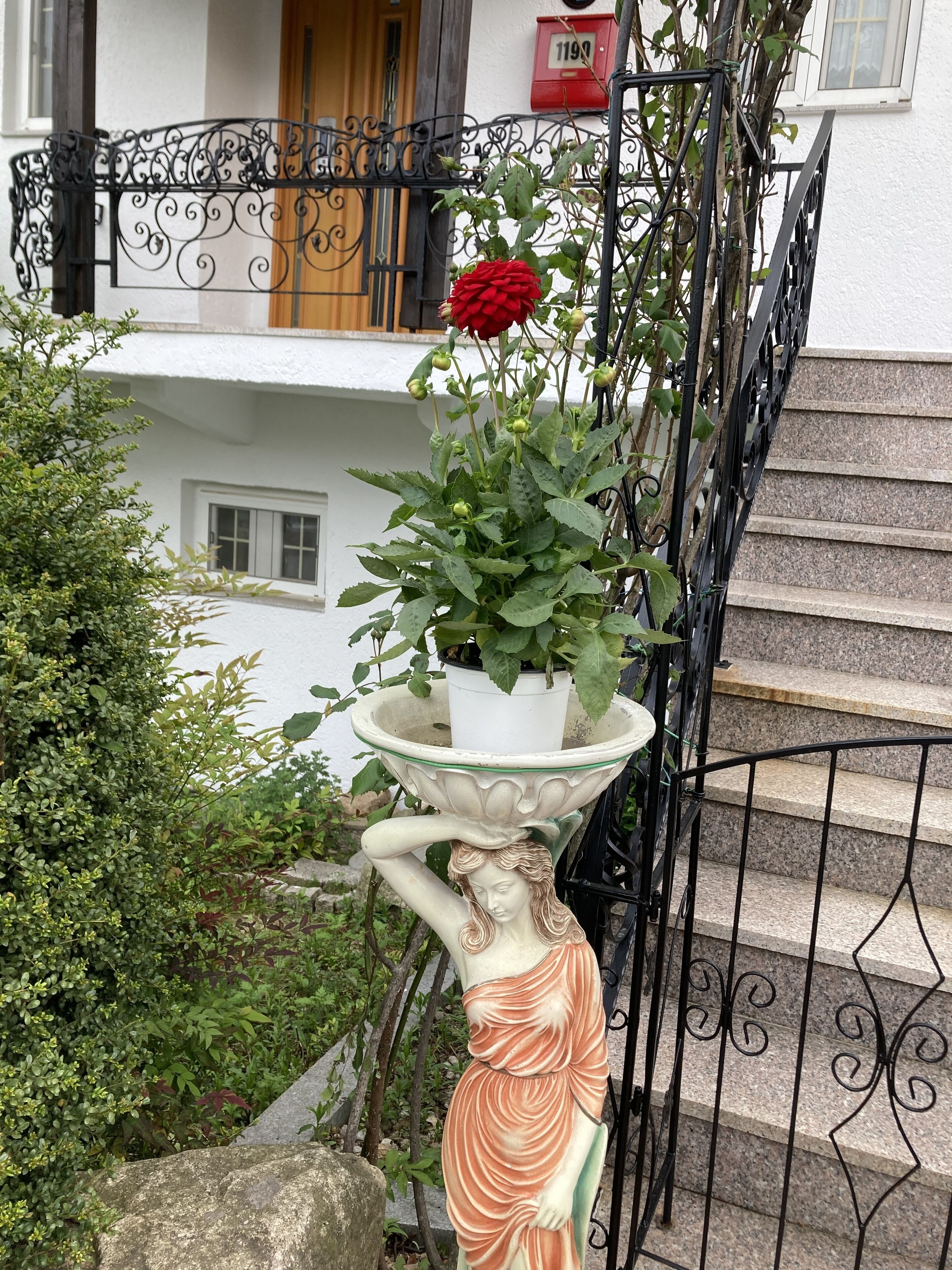 A statue of a woman holding a flower, a potted plant with a red rose, and a staircase with a black railing.