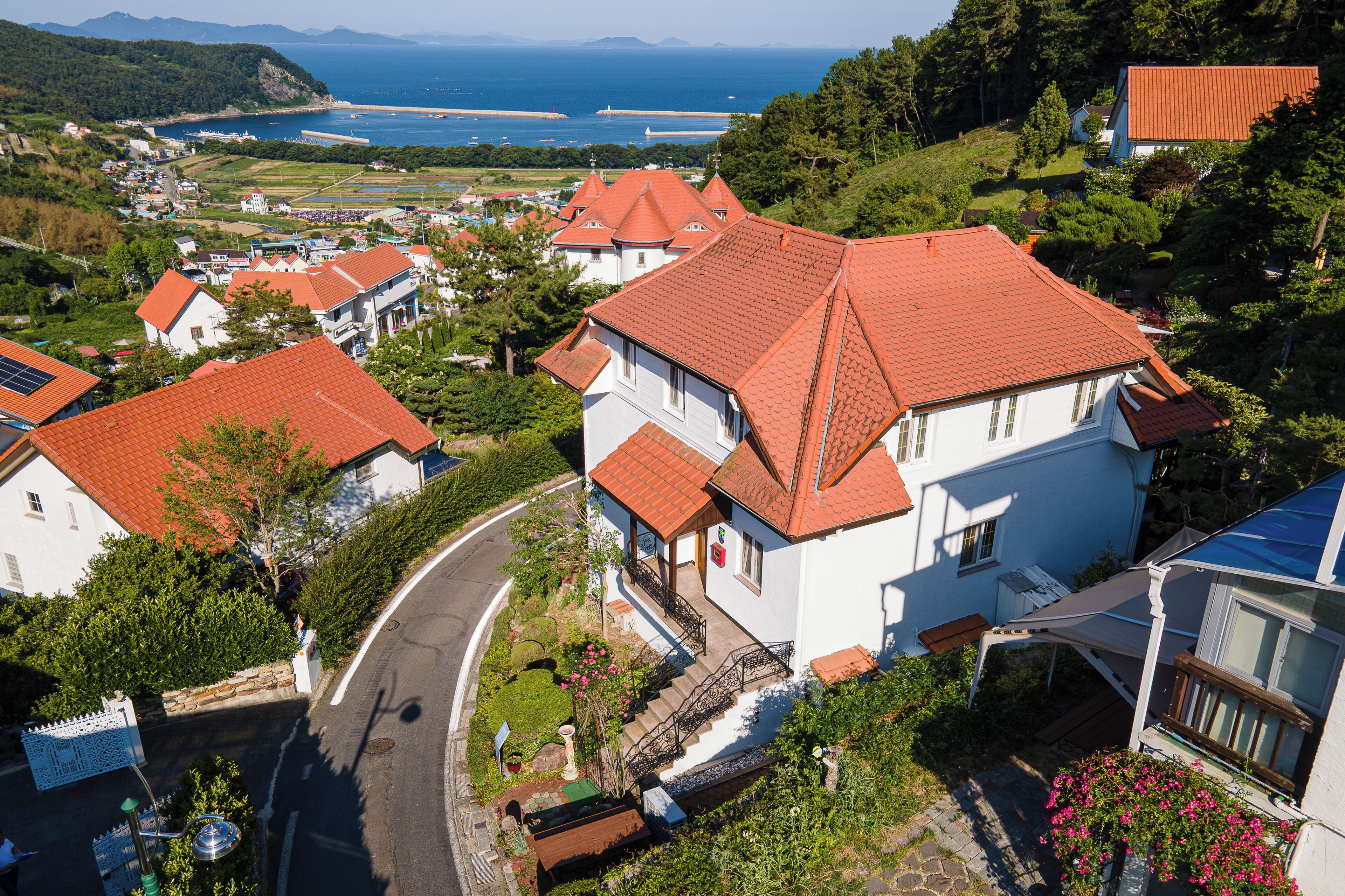 A residential area with houses featuring red-tiled roofs, a winding road, and a view of the sea and hills in the background.