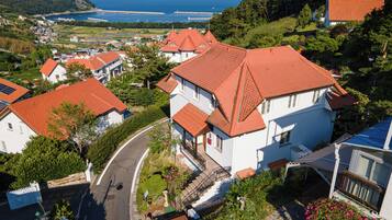 A residential area with houses featuring red-tiled roofs, a winding road, and a view of the sea and hills in the background.