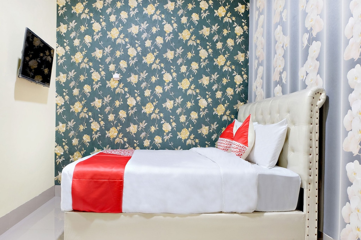 Una cama con una colcha roja y blanca, un cabecero capitonado y almohadas blancas. La habitación tiene papel tapiz floral y una cortina con flores blancas.