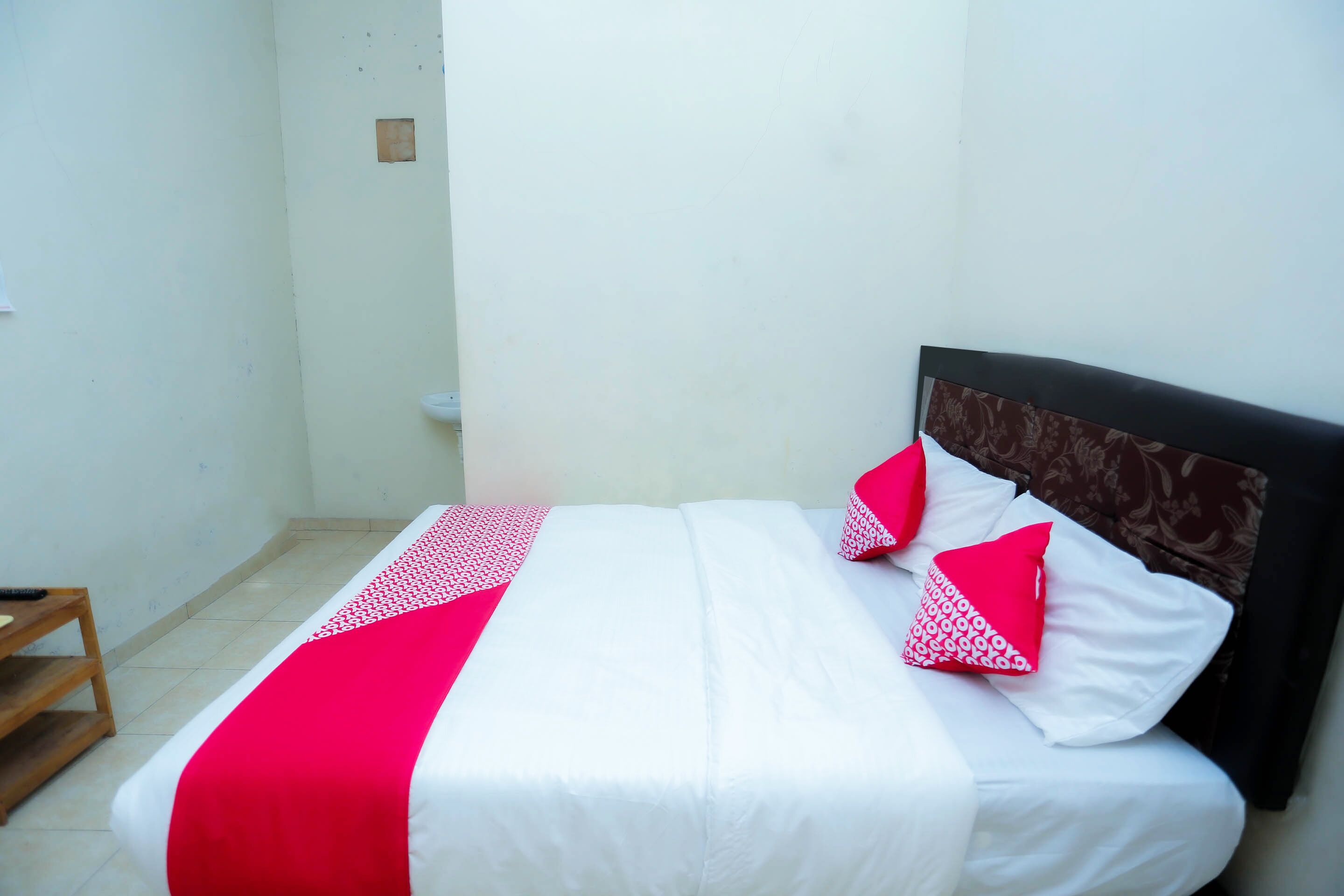 A neatly made bed with a white and red bedspread, a wooden nightstand, and a plain white wall.
