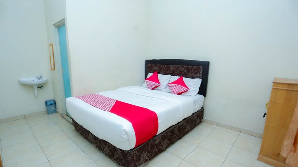 A neatly made bed with a white and red bedspread, a wooden headboard, and a small bedside table with a lamp. - OYO 2692 Penginapan Mba Ros (Banjarbaru)