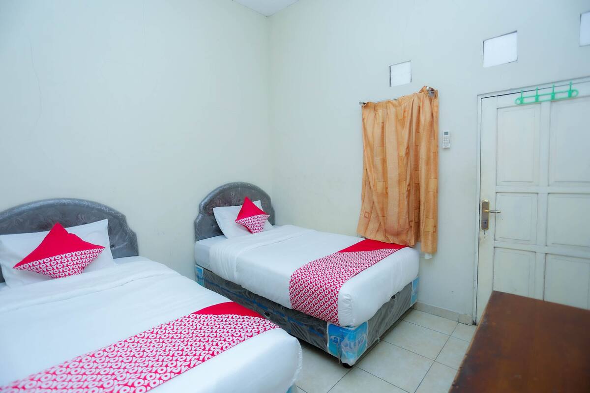Quarto com duas camas, cada uma com roupa de cama branca e detalhes vermelhos, uma cabeceira de madeira e uma porta com suporte para toalhas verde.