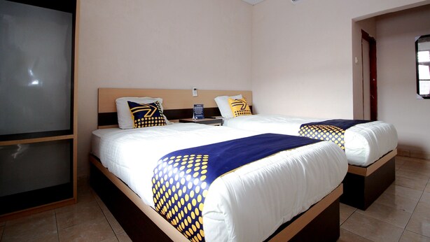 Une chambre d’hôtel avec deux lits, chacun doté d’une tête de lit en bois, de draps blancs et de plaids aux motifs bleus et jaunes. On y trouve une table de chevet avec une lampe et une pancarte indiquant le type de chambre.