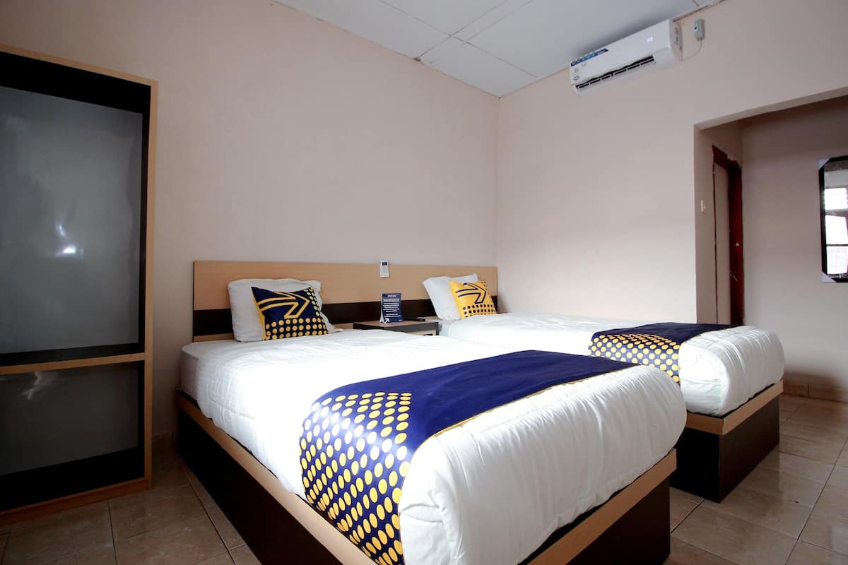 A hotel room with two beds, each with a wooden headboard, white bedding, and blue and yellow patterned throw blankets. There is a nightstand with a lamp and a sign indicating the room type.